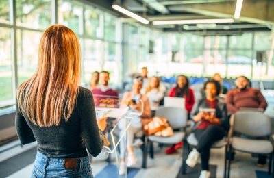 A woman presenting content at a workshop to a group of people sitting in an open plan conference room