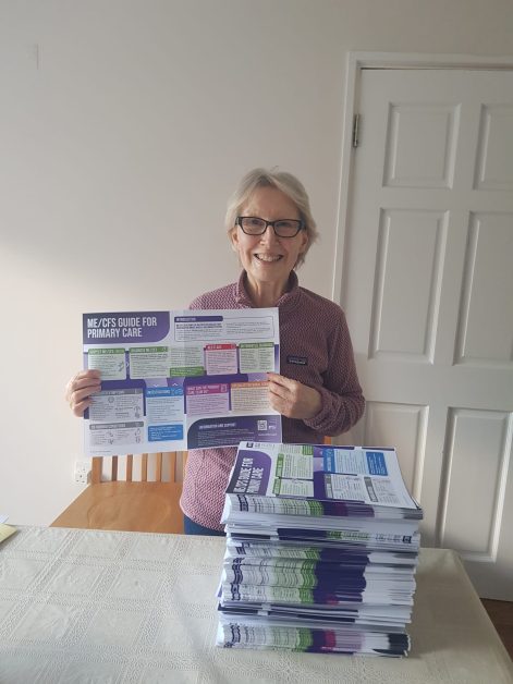 Irish-MECFS-Association -Margaret-Nolan IMAGE DESCRIPTION: Photo of a woman in her home, holding up a copy of the MEA Primary Care Infographic Poster