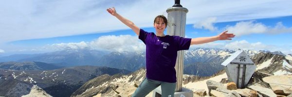 Image of Fundraiser Isabel on her climb to Mount Aneto, the highest mountain in the Pyrenees. She is standing with arms outstretched and wearing a purple MEA tshirt.