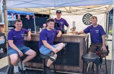 We’re Team The Flying Bulls. With the race in London, we’re building a true British classic: a pub – complete with working taps and proper snacks to make the judges feel at home and the crowd feel thirsty. We’re also racing for charity, supporting friends affected by ME/CFS and its impact on mental health. For us, the pub is about connection, laughter and bringing people together for a good chat – this time at full speed down the hill. Photo of the team wearing ME Association tshirts sitting in their outdoor bar.