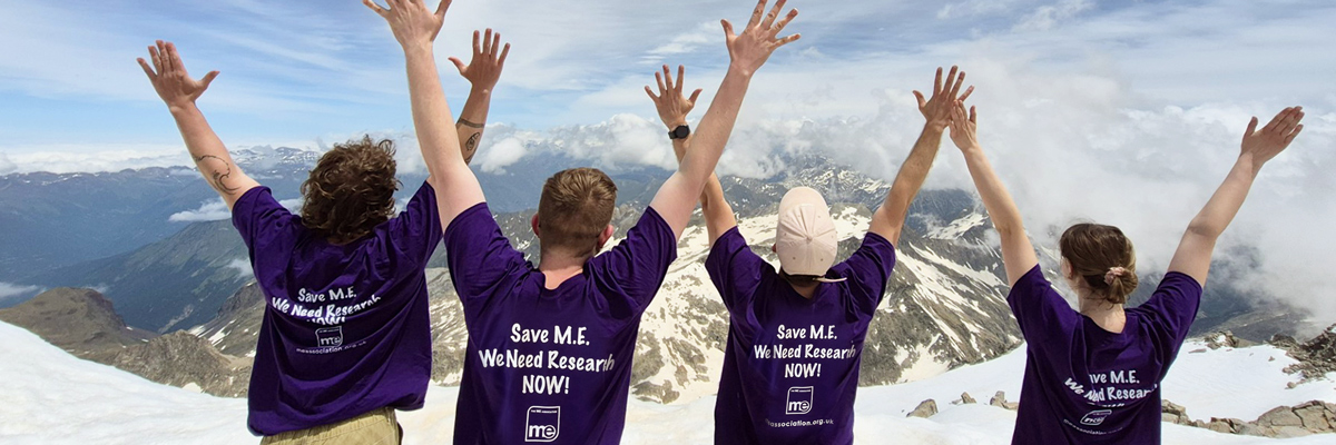 Group challenge showing 4 people at Aneto Mountain peak in Spain, with their arms in the arms in the air and their backs to the camera showing ME Association tshirts with the wording "Save M.E. We need research now. ME Association"