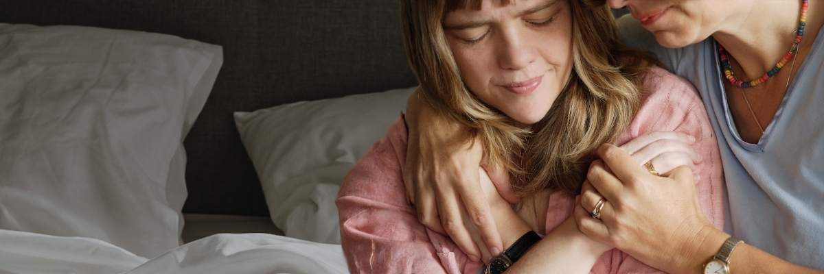 Picture of a woman sick in bed with her eyes closed, being hugged by a comforting family member